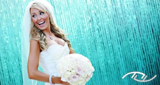Celena smiling in her stunning bridal gown and holding her beautiful bouquet while standing in front of the blue fountain at the Surf & Sand Hotel in Laguna Beach, CA. (Photo Credit: Jim Kennedy Photographers)